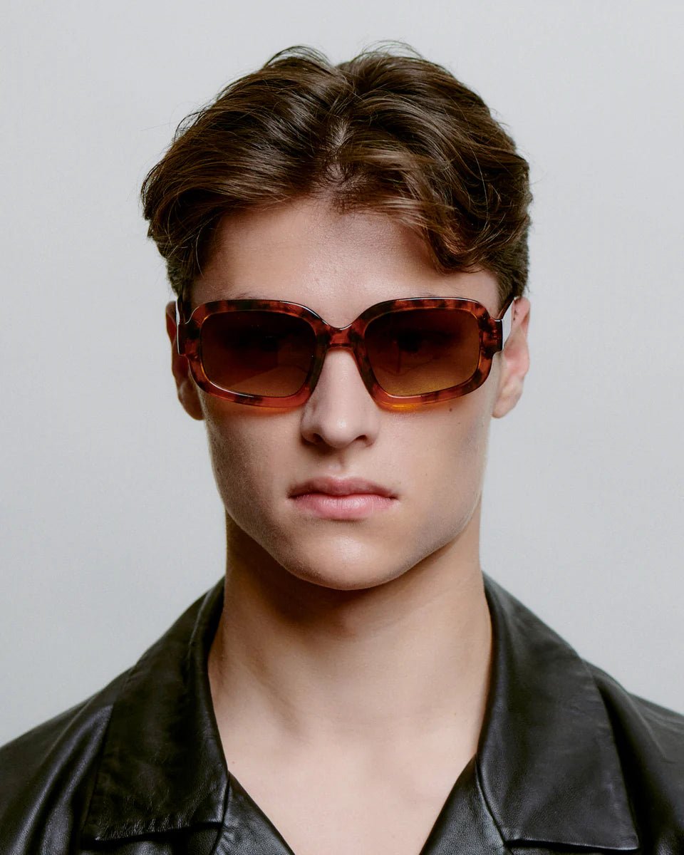 Havana-style Pluto sunglasses by A. Kjaerbede with round frames and dark tinted lenses, resting on a white surface.