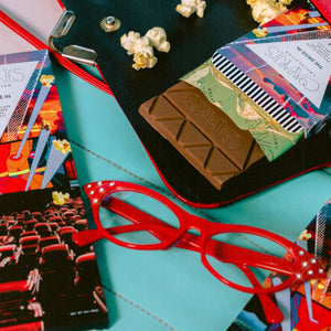 Close-up of a turquoise surface with red glasses, popcorn, chocolate, and movie tickets.