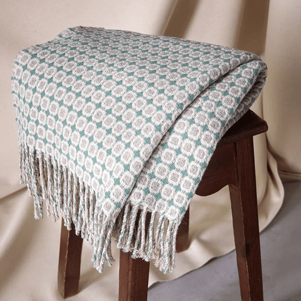 Patterned throw blanket with fringes draped over a wooden stool against a neutral background