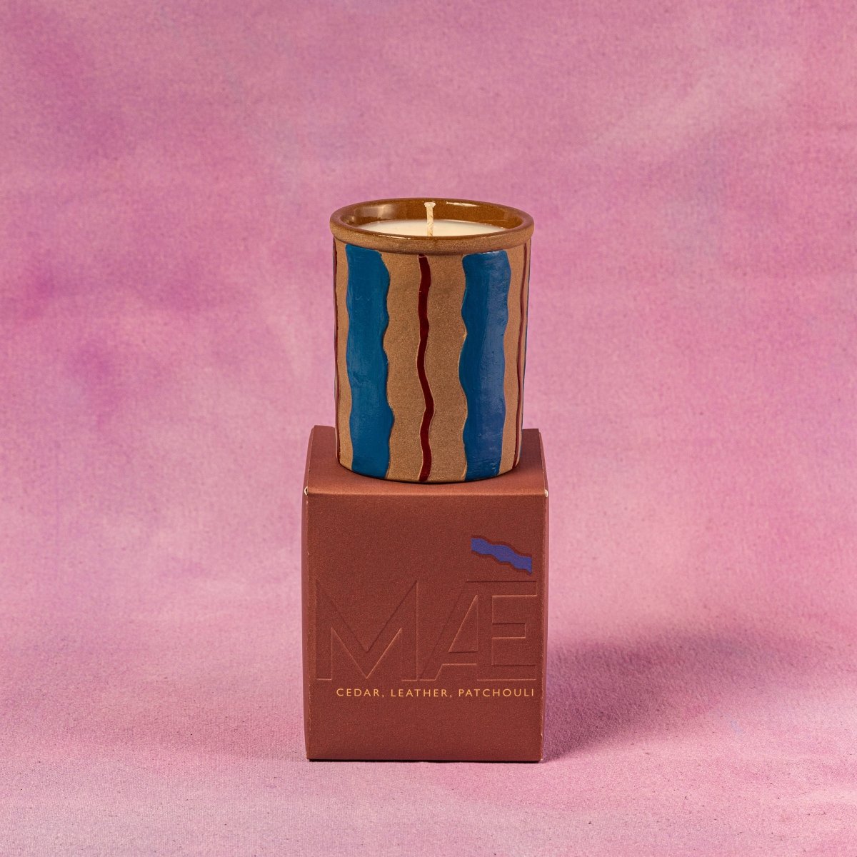 Candle in a wooden container on top of a brown box with 'MVE' branding against a pink background