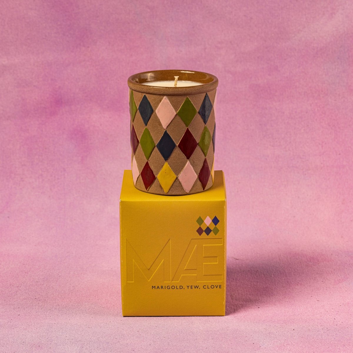 Candle in a patterned container on top of a yellow box with a pink background